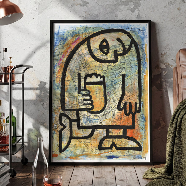 A framed abstract art print featuring a black line-drawn figure on a textured blue, yellow, and orange background, displayed in a cozy, rustic living room setting next to a leather armchair, a copper lamp, and a wooden floor.