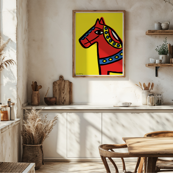 A framed art print featuring a stylized red horse on a bright yellow background, hanging on a rustic white wall in a sunlit kitchen with wooden shelves, a table, and dried grasses.