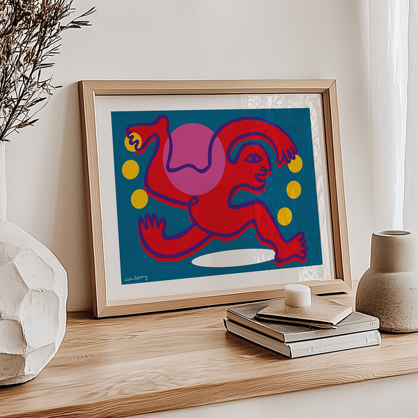 A framed abstract art print featuring a red figure and yellow circles on a blue background, styled on a wooden console table with books, a candle, and ceramic vases.